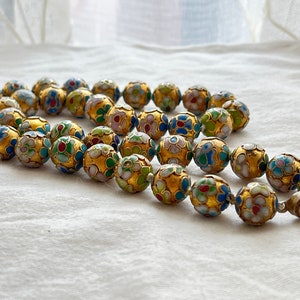 May include: A gold necklace with round beads decorated with colorful enamel flowers. The beads are strung on a silver chain with a clasp.