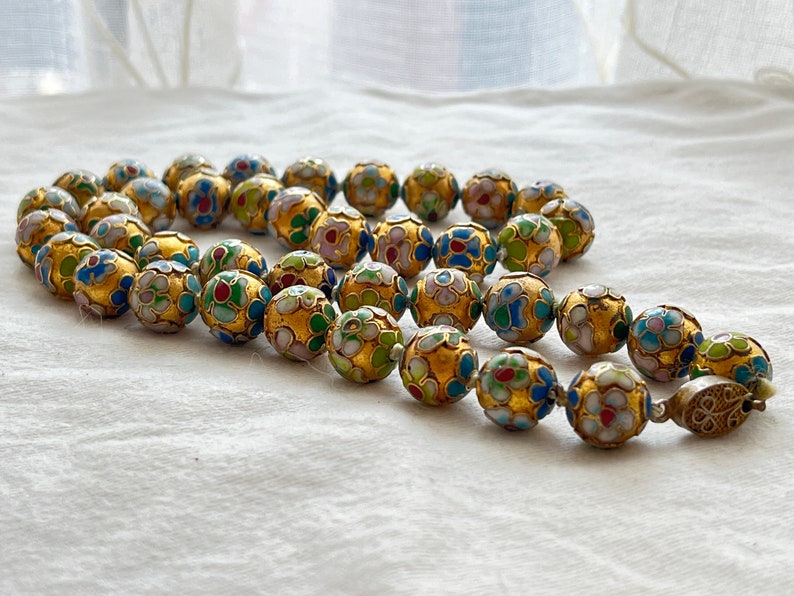 May include: A gold necklace with round beads decorated with colorful enamel flowers. The beads are strung on a silver chain with a clasp.