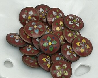 May include: A pile of 16 round, red, enamel buttons with a white flower design and colorful accents. The buttons are arranged in a random pattern on a white surface.