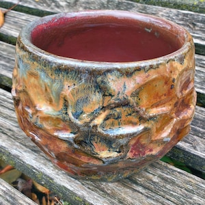 May include: A handmade ceramic bowl with a textured exterior and a rich color palette. The bowl features a reddish-brown interior and a multi-toned exterior with shades of orange, brown, blue, and hints of white. The bowl is sitting on a wooden surface.