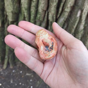 May include: A small, irregularly shaped, ceramic sculpture with a reddish-orange glaze and black accents. The sculpture has a smooth, rounded surface and a slightly textured finish.