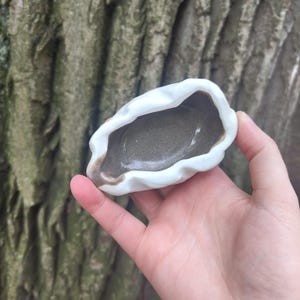 May include: A small, handmade ceramic dish with a unique, organic shape. The dish features a white exterior with a rippled edge and a gray interior. A touch of brown accents the rim, adding a natural aesthetic. Perfect for holding small items.