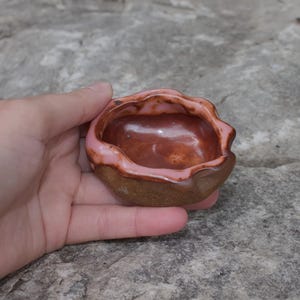 May include: A small, hand-held ceramic bowl with an irregular, wavy rim. The interior features a glossy, deep reddish-brown glaze, while the exterior has a textured, earthy brown finish. A soft pink glaze accents the top edge of the wavy rim.