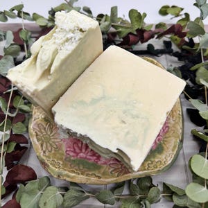 Puede incluir: Dos jabones hechos a mano, uno con la parte superior texturizada, descansan sobre un plato de cerámica decorativo con un diseño floral. Los jabones son de color verde claro y blanco, con un efecto veteado en uno. Hojas de eucalipto rodean el plato.
