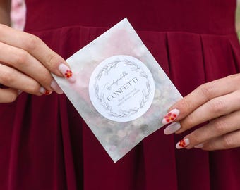 Può includere: Un piccolo pacchetto quadrato di coriandoli biodegradabili, fatti con petali di fiori secchi, tenuto contro un vestito bordeaux. Il pacchetto ha un'etichetta circolare bianca con la parola "Confetti" e un design a ghirlanda floreale.