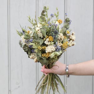 May include: A bouquet of dried flowers featuring muted green eucalyptus leaves, small clusters of yellow billy buttons, white yarrow, and sprigs of blue thistle. The stems are gathered together and tied with a thin piece of natural twine.