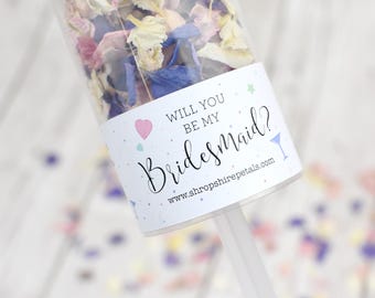 Könnte beinhalten: Ein weißer Konfetti-Kanone mit einem Etikett, auf dem "Willst du meine Brautjungfer sein?" steht, mit einem rosa Herzen, blauen Konfetti und einem Martini-Glas-Symbol. Die Website www.shropshirepetals.com ist auf dem Etikett aufgedruckt.