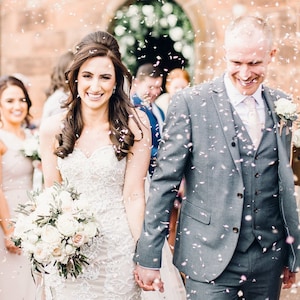 May include: A bride and groom walking down the aisle after their wedding ceremony, showered with confetti. The bride is wearing a white lace wedding dress and carrying a bouquet of white and pink flowers. The groom is wearing a gray suit with a pink tie.