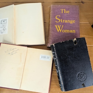 May include: Four vintage books are displayed on a wooden surface. One book is open, revealing blank pages and a small illustration. Another book has a purple cover with the title "The Strange Woman" in yellow. A third book is open with handwritten text.