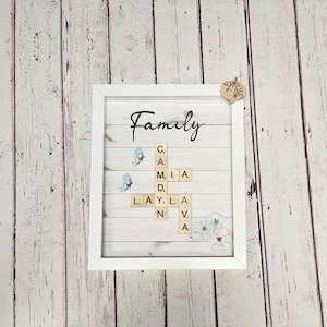 May include: A white framed picture with a woodgrain background. The word "Family" is written in black cursive script. The word is spelled out using letter tiles in a crossword puzzle format. The background is decorated with blue butterflies and white flowers.