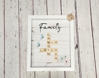 May include: A white framed picture with a woodgrain background. The word "Family" is written in black cursive script. The word is spelled out using letter tiles in a crossword puzzle format. The background is decorated with blue butterflies and white flowers.