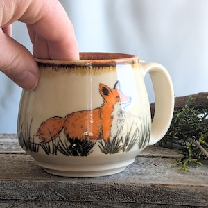May include: Cream-colored ceramic mug with a brown rim and handle. The mug features a hand-painted orange fox illustration with black and green grass. The mug is held by a hand.
