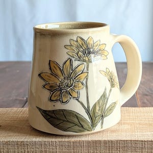 以下が含まれることがあります： クリーム色のセラミックマグカップ。大きなハンドルが付いています。黄色いヒマワリと緑の葉と茎の手描きのデザインが特徴です。木製の表面に置かれています。