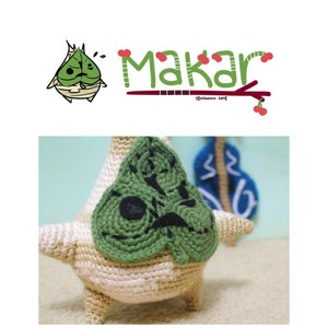 Może przedstawiać: Pluszowa zabawka w kształcie liścia, wykonana na szydełku, z zieloną twarzą i czarnymi oczami. Zabawka stoi na beżowej powierzchni. W tle widoczny jest tekst "Makar".