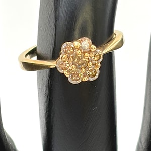 May include: A gold ring featuring a flower-shaped cluster setting. The center of the flower is adorned with seven small, light brown, round-cut stones arranged in a circular pattern, held in place by small gold prongs on a smooth gold band.