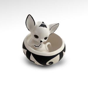 May include: A white ceramic chihuahua figurine with black accents, sitting inside a black and white patterned bowl. The chihuahua has black-tipped ears and a small black triangle on its head. The bowl has a geometric design.
