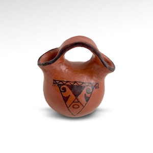 May include: A small, hand-crafted terracotta pottery jug with a unique design. The jug has a wide, rounded body and a narrow neck with a handle. The jug is decorated with a black geometric pattern.