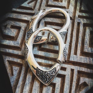 Può includere: Due orecchini a cerchio d'argento con intricati disegni floreali. Gli orecchini sono su una superficie di legno con un motivo geometrico.