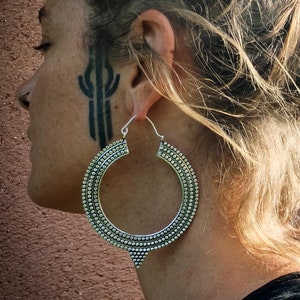 Peut inclure: Une paire de grandes boucles d'oreilles en argent avec un design texturé et perlé. Les boucles d'oreilles sont portées par une personne aux cheveux blonds.