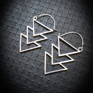 Peut inclure: Une paire de boucles d'oreilles créoles en argent avec un motif géométrique composé de trois triangles superposés.