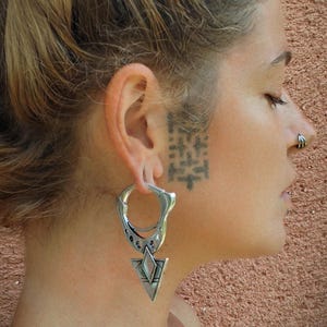 Puede incluir: Pendientes geométricos plateados con un colgante triangular. La persona que los lleva tiene un piercing en la nariz y un tatuaje en la cara. La persona tiene el pelo recogido en un moño. El fondo es de color marrón rojizo.