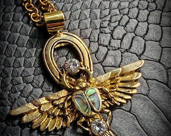 May include: A gold pendant necklace featuring an ankh symbol with a scarab beetle design. The ankh is adorned with clear gemstones and the scarab beetle has a green abalone shell.