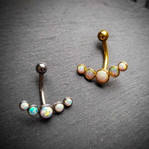 Peut inclure: Deux piercings de nombril. L'un est argenté, l'autre doré. Les deux ont une barre incurvée avec une petite boule au sommet et une rangée de pierres opalescentes. Les pierres sont disposées en demi-cercle.