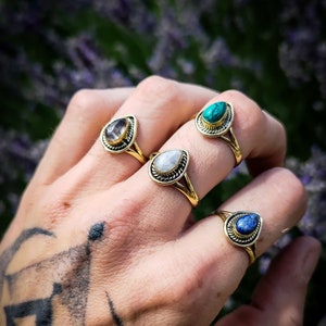 Puede incluir: Cuatro anillos de oro con diferentes piedras preciosas de color en forma de lágrima. Los anillos se llevan en una mano con un tatuaje.