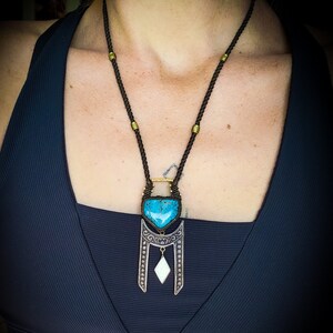 Peut inclure: Un collier avec un pendentif en pierre turquoise, un cadre orné argenté et un charme blanc en forme de diamant suspendu en dessous. Le collier est fait d'un cordon noir et de perles dorées.