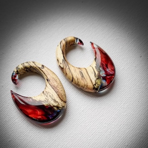 Puede incluir: Un par de dilatadores de oreja de madera y resina con un esquema de color rojo, blanco y azul. Los dilatadores tienen forma de media luna y un acabado liso y pulido.