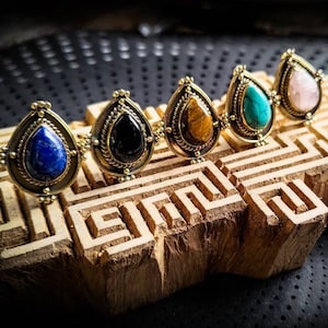 Könnte beinhalten: Fünf goldene Ringe mit tropfenförmigen Edelsteinen. Die Steine sind Lapislazuli, schwarzer Onyx, Tigerauge, Türkis und Rosenquarz.