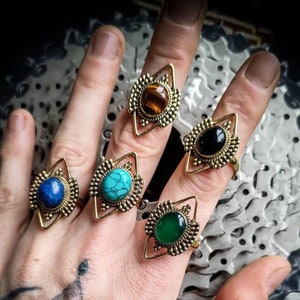 Peut inclure: Cinq bagues en or avec des pierres précieuses de différentes couleurs. Les bagues présentent un design géométrique avec un sommet pointu et un fond arrondi. Les pierres précieuses sont turquoise, verte, noire, brune et bleue.