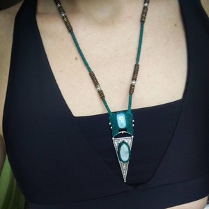 Peut inclure: Une femme portant un soutien-gorge de sport noir et un pantalon gris avec un collier à pendentif vert et argent. Le pendentif présente un design triangulaire avec deux pierres ovales.