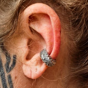 May include: A silver-toned ear cuff featuring a textured, scale-like pattern. The jewelry piece is designed to wrap around the outer cartilage of the ear, showcasing a detailed, bumpy surface finish that covers the entire exterior of the circular band.
