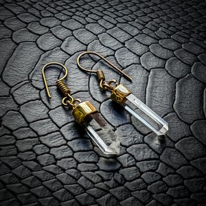 Peut inclure: Une paire de boucles d'oreilles en cristal de quartz clair avec des accents en métal doré. Les boucles d'oreilles présentent un design à crochet et sont posées sur un fond noir texturé. Les cristaux mesurent environ 2,5 cm de long.
