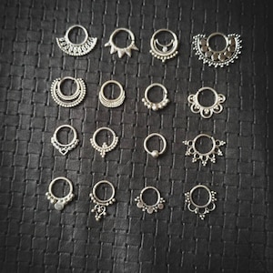 Puede incluir: Una colección de 18 anillos de nariz de plata con varios diseños decorativos, que incluyen formas geométricas, cuentas y filigrana.