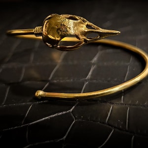 Puede incluir: Un brazalete dorado con un diseño de cráneo de pájaro. El brazalete está abierto y tiene una forma simple y curvada.