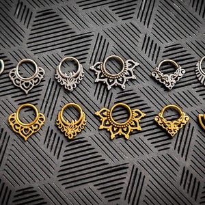 Puede incluir: Una variedad de pendientes y anillos de nariz decorativos en tonos plateados y dorados. La joyería presenta varios diseños intrincados, incluyendo patrones florales y geométricos. Las piezas están dispuestas sobre una superficie texturizada de color gris oscuro.
