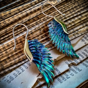May include: A pair of iridescent blue and green wing-shaped earrings with gold accents. The earrings are hanging from silver hooks.
