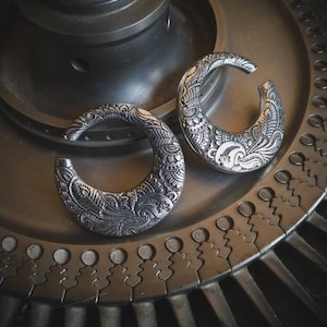 Può includere: Due dilatatori per orecchie in argento con intricati disegni floreali. I dilatatori hanno la forma di una mezzaluna e presentano un motivo dettagliato e ornato.