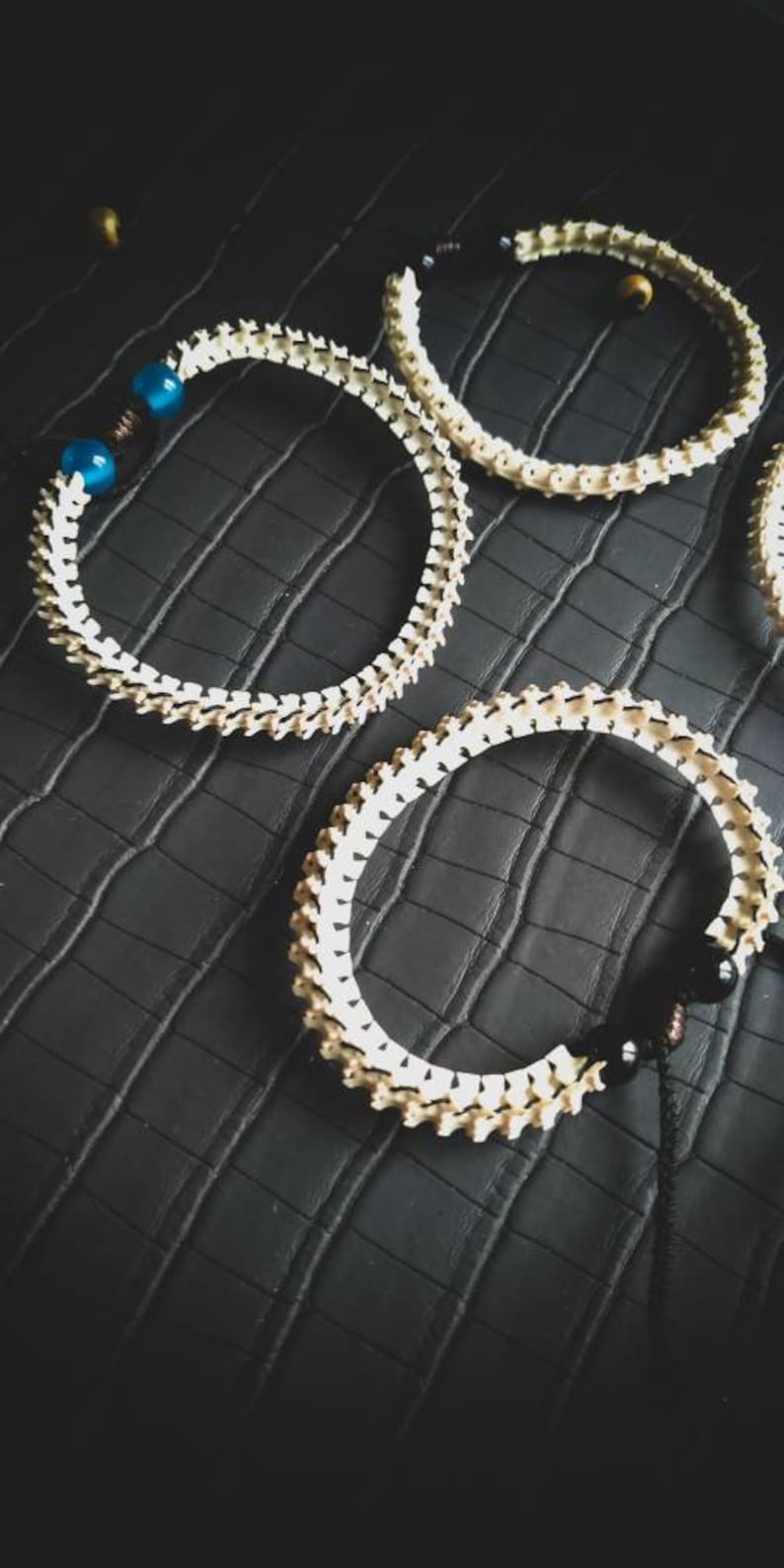 Peut inclure: Trois bracelets en os blanc avec des perles noires et bleues. Les bracelets sont sur une surface textur&eacute;e noire.