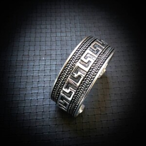 Peut inclure: Un bracelet manchette argenté avec un motif de clé grecque. Le bracelet présente une bordure tressée et un motif géométrique détaillé. Le métal a une finition polie et le bracelet est présenté sur une surface noire texturée.
