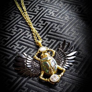 Può includere: Una collana con ciondolo in oro a forma di scarabeo. Lo scarabeo è realizzato in madreperla di abalone iridescente e ha ali nere. La collana è su uno sfondo nero con un motivo geometrico.