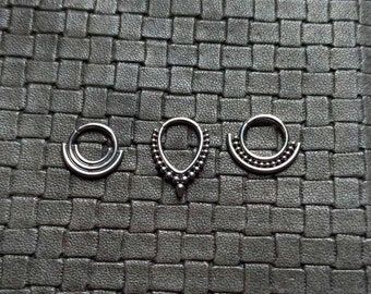 Peut inclure: Trois anneaux de septum argentés avec des designs différents. La première bague a un design simple à double boucle. La deuxième bague a une forme de larme avec un bord perlé. La troisième bague a un bord perlé et un design incurvé en demi-cercle.