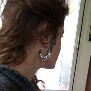 Peut inclure: Une boucle d'oreille en argent avec un motif décoratif. La boucle d'oreille est portée à l'oreille gauche d'une personne aux cheveux bruns.