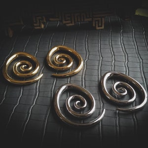 Op de afbeelding: Twee paar spiraalvormige oorpluggen, één paar is goud en het andere is zilver. De pluggen liggen op een zwart, getextureerd oppervlak.