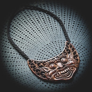 May include: A wooden necklace with a carved, detailed, brown, demonic face pendant. The pendant is shaped like a crescent moon and has a black cord.