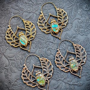 Peut inclure: Quatre boucles d'oreilles créoles en laiton, chacune présentant un motif de scarabée central incrusté de nacre d'ormeau irisée. Le scarabée est flanqué de deux ailes stylisées en forme de feuille avec des détails découpés, suspendues à un anneau.