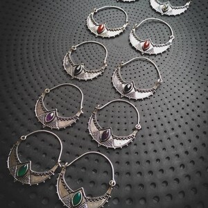 Peut inclure: Un ensemble de dix boucles d'oreilles créoles en argent avec des pierres précieuses de différentes couleurs. Les boucles d'oreilles sont en forme de croissant et présentent des détails complexes. Les pierres précieuses sont vertes, rouges, violettes et noires.