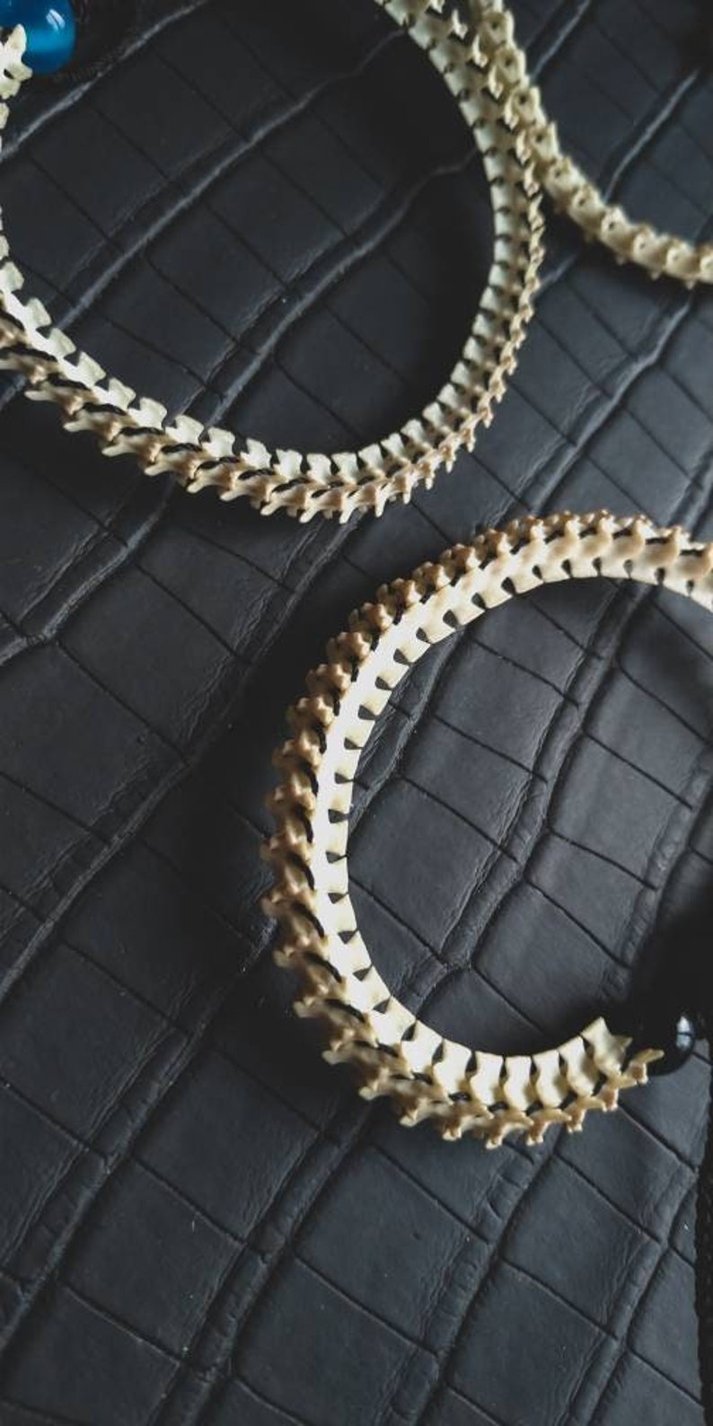 Peut inclure: Deux colliers en os sur une surface textur&eacute;e noire. Les colliers sont faits de petites pi&egrave;ces d'os blanches qui sont reli&eacute;es entre elles pour former un cercle. Les colliers sont pos&eacute;s &agrave; plat sur la surface.